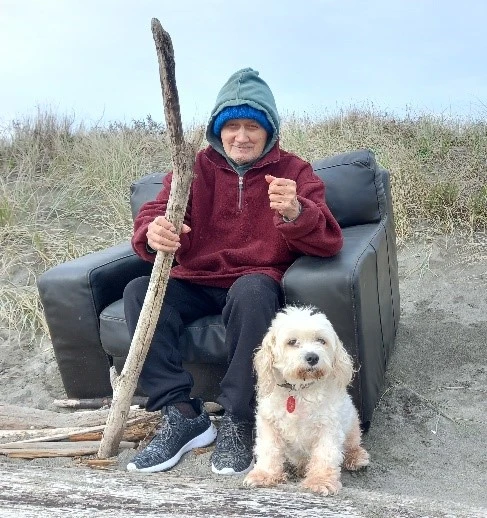 A koroua (older male) sitting on the beach, holding a tree branch and accompanied by his small white dog